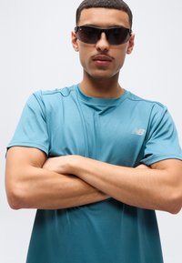 Νέος άνδρας που φοράει μαύρα γυαλιά ηλίου και μπλουζάκι αθλητικό New Balance σε τιρκουάζ χρώμα, με σταυρωμένα τα χέρια, μπροστά σε απλό φωτεινό φόντο.