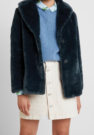 Person iført mørk navy kunstpelsfrakke over lyseblå striktrøje og hvid knap-foran nederdel med synlig blomsterprint på skjortekrave og manchetter.