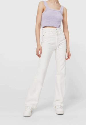 Kobieta w miękkim fioletowym krótkim topie, białych wysokich dżinsach i białych trampkach, stojąca na tle jednolitego jasnego tła.