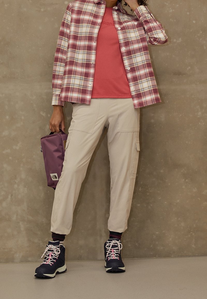 Personne portant un pantalon cargo beige, une chemise à carreaux rouge par-dessus un t-shirt rouge, des chaussures de randonnée bleu marine, et tenant un sac violet contre un mur uni.