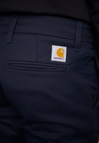 Navyblaue Baumwollhosen mit glattem Textil, ausgestattet mit einer Gesäßtasche und einem weißen Etikett mit einem orangefarbenen Logo.