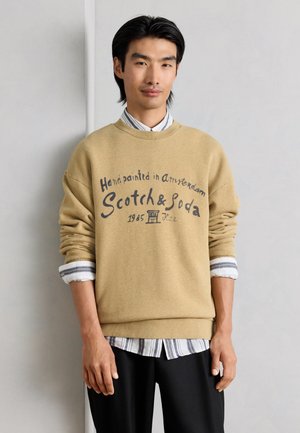 Άνδρας φοράει μπεζ φούτερ με το κείμενο "Handpainted in Amsterdam Scotch & Soda 1985" πάνω από ριγέ πουκάμισο και μαύρο παντελόνι, στέκεται μπροστά σε γκρίζο τοίχο.