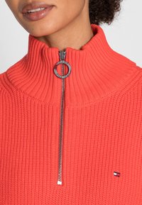 Korallfärgad stickad tröja med hög ribbad krage och silverfärgad dragkedja. Har en liten logotypdetalj på vänster sida. Texturerat tyg.