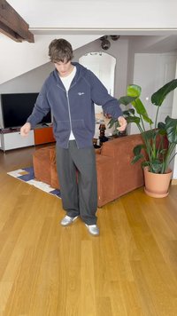 Sweatshirt azul marinho com capuz, fecho éclair e bolsos, combinado com calças soltas cinzas escuras e ténis prateados, de pé numa sala de estar.