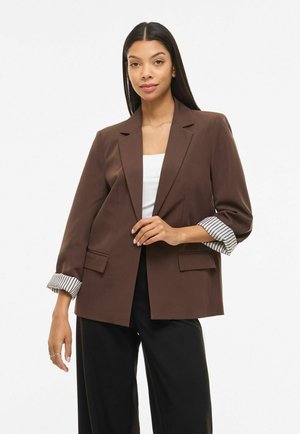 Vrouw draagt een bruine blazer met opgerolde, gestreepte mouwen over een wit shirt en zwarte broek, staande tegen een effen witte achtergrond.