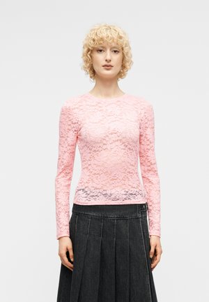 Asmuo dėvi ilgomis rankovėmis rožinę nėriniuotą palaidinę ir aukštu liemeniu tamsią klostuotą sijoną, stovintis priešais paprastą šviesią foną.