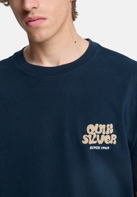 Μπλε μπλουζάκι από βαμβάκι με στρογγυλή λαιμόκοψη, με μπεζ λογότυπο γραφικού χαρακτήρα στο αριστερό στήθος που γράφει «QUIKSILVER SINCE 1969.»