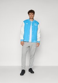 Blue varsity jacket with white sleeves, logo on chest, gray sweatpants with white side stripes, and black sneakers.
