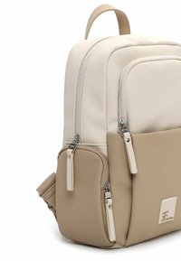 Beżowo-kremowy plecak wykonany z materiału syntetycznego, wyposażony w wiele kieszeni na zamek, górny uchwyt oraz logo na przodzie.
