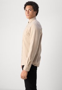 Camicia beige chiaro a maniche lunghe con colletto a bottoni, due tasche sul petto e un fit rilassato, abbinata a pantaloni neri. Tessuto texture.
