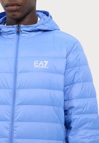 Jachetă pufoasă de culoare albastră deschisă, cu glugă, având închidere prin zip în față și cusături diagonale. Logo-ul EA7 brodat pe piept.