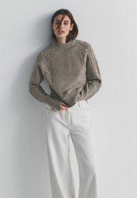 Gebreide grijze trui met een hoge kraag en kabelgebreid patroon, gecombineerd met losse witte broek. Zachte textuur, ontspannen pasvorm en lange mouwen.