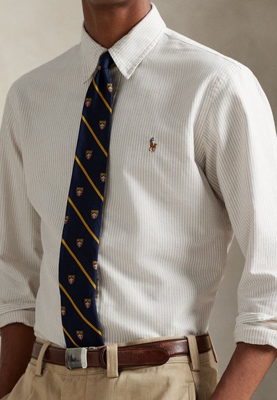 Vyras dėvi smėlio spalvos dryžuotą marškinių su sagomis ir užriestomis rankovėmis, tamsiai mėlyną kaklaraištį su geltonomis juostomis ir herbo ženklais, rudą odinį diržą ir chaki spalvos kelnes.