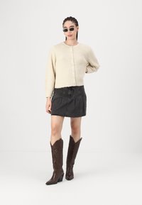 Cardigan di colore crema, con texture e bottoni; abbinato a una gonna di denim scuro e stivali da cowboy in pelle marrone con cuciture decorative.