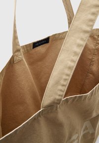 Tote-Bag aus hellbraunem Stoff mit auffälligen Nähten. Innen hat sie eine glatte Textur und zeigt das Markenschild in Schwarz mit weißer Schrift.