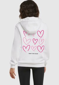 Felpa bianca con più cuori rosa sulla schiena e la scritta "FIDATI DEL TUO CUORE" in nero. Tessuto morbido, maniche lunghe e cappuccio.