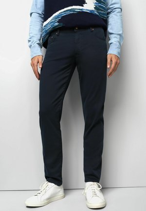 Mann in dunkelblauen Slim-Fit-Hosen, weißen Sneakers und einem hellblauen Hemd unter einem gemusterten Pullover, stehend vor einer weißen Wand.