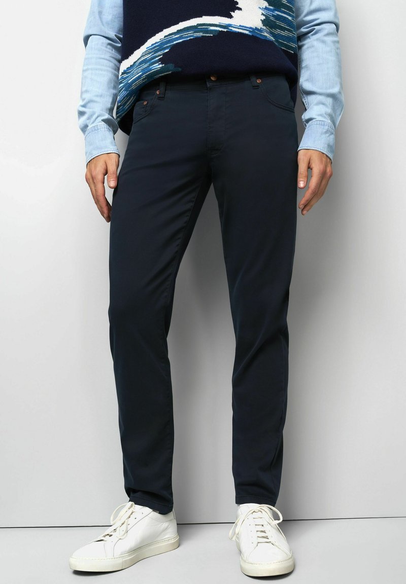 Homme portant un pantalon slim-fit marine, des baskets blanches et une chemise bleu clair sous un pull à motifs, se tenant devant un mur blanc.