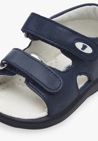Sandale en cuir bleu marine avec deux sangles réglables en Velcro, doublure intérieure blanche et semelle rembourrée. Présente des découpes pour la respirabilité.