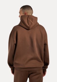 Sweat à capuche marron au fit décontracté, avec des poignets et un ourlet côtelés. Texture lisse avec un design simple, vu de l'arrière.