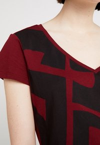 Burgundy bomullströja med en svart abstrakt design, korta ärmar och V-hals. Slät textur med en avslappnad passform.