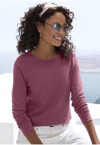 Rosa strukturierter Langarmpullover mit Rautenmuster, Rundhalsausschnitt und umgeschlagenen Bündchen, kombiniert mit weißen Hosen und Sonnenbrille.