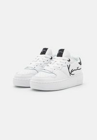 Hvide læder sneakers med en tyk gummisål, snørebåndsdesign, perforeret tåboks og sorte logo aksenter på tungen og siden.