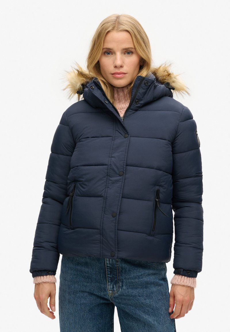 Veste matelassée bleu marine avec capuche et bord en fausse fourrure, dotée d'une poche zippée, de boutons-pression et d'une texture matelassée. Portée sur un pull rose.
