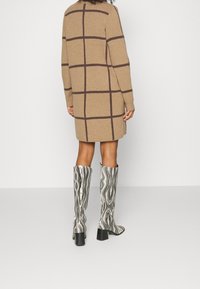 Una persona che indossa un cappotto a scacchi marrone e stivali a fantasia fino al ginocchio, girata di spalle contro uno sfondo bianco semplice.