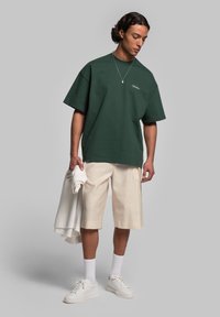 Grön oversized t-shirt med korta ärmar, kombinerad med krämfärgade shorts. Vita sneakers och en ljus sweatshirt hängande över armen.