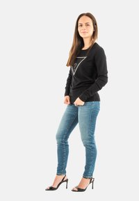 Sudadera negra de manga larga con un diseño triangular plateado y detalles de tachuelas, combinada con unos jeans ajustados azules y sandalias de tacón negro.