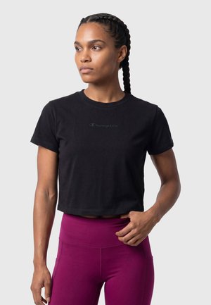 Vrouw met gevlochten haar die een zwart cropped T-shirt en paarse leggings met hoge taille draagt, kijkt iets opzij.