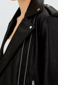 Czarna skórzana kurtka motocyklowa z szerokim kołnierzem, zapięciem na zamek błyskawiczny z przodu, metalowymi ćwiekami i kieszeniami na zamek. Gładka tekstura z srebrnymi akcentami.