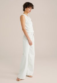 Fehér ujjatlan felső drapált nyakkal, magas derekú, fehér, bő szárú farmernadrággal párosítva. Sima textúra és minimalista design.