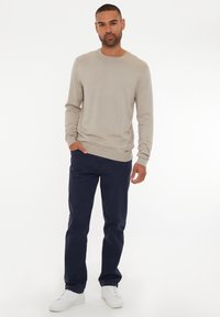 Beiger Pullover mit langen Ärmeln und Rundhalsausschnitt; dunkelblaue gerade Hosen; weiße Sneakers; glatte Textur, lässige Passform.