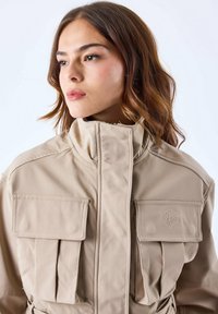 Beige, textuurjacket met een hoge kraag, twee grote borstzakken en een geborduurde logo-detail. De buitenkant heeft een gestructureerd ontwerp.