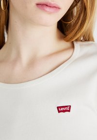 Fehér pamut póló, amelyen bal oldalon, a mellkasnál piros hímzett Levi's logó található. A nyakrésze kerek, látható varrással.