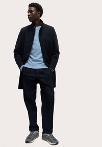 Mørk navyfrakke over en lyseblå sweater, kombineret med mørke bukser og grå sneakers. Frakken har en høj krave og knapper.