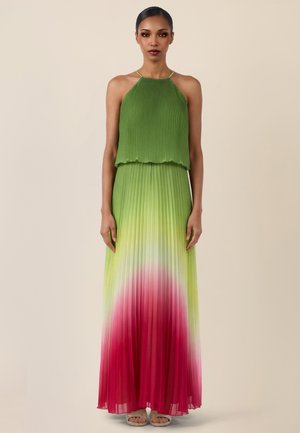 Kobieta w zielonej plisowanej sukience na szyję bez rękawów, która przechodzi w różowy kolor przy brzegu, stojąca na tle jednolitego beżowego tła.