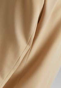 Beżowy poliester o gładkiej teksturze, z delikatnym fałdowaniem i miękkim układaniem. Główny nacisk kładziony jest na subtelny połysk materiału i jego złożenia.