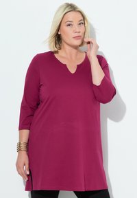 Fuksia tunika ylin, jossa on 3/4 hihat, V-muotoinen kaula-aukko ja sivurei'ät; valmistettu pehmeästä, joustavasta kankaasta, suunniteltu mukavuutta varten.