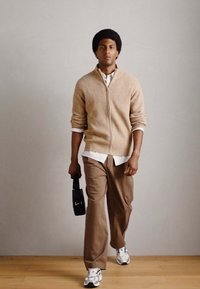 Beige strikket zip-up genser over hvit skjorte, brune vide bukser og hvite sneakers. Holder en svart bag. Enkel bakgrunn.