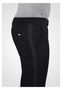 Zwarte broek met een grijze streep langs de zijkant, met een gestructureerde tailleband en een klein zakje met drie decoratieve knopen.