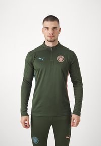 Zöld sportos felső, cipzáros gallérral, kék Puma logóval és Manchester City emblémával. Hosszú ujjú és mintás szövetkialakítás.