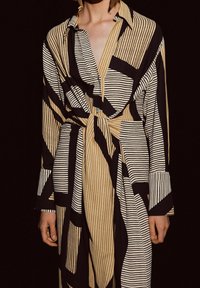 Eine Frau trägt ein langes, gestreiftes Kleid mit schwarzen, beigen und weißen geometrischen Mustern sowie einem Knoten in der Taille vor einem dunklen Hintergrund.