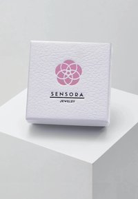 Bela teksturirana škatla za nakit s rožnatim logotipom in črnim besedilom "SENSORA JEWELRY," postavljena na svetlo sivo površino.