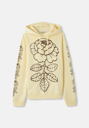 Sudadera con capucha amarilla que presenta un gran diseño floral marrón en la parte frontal y hojas estilizadas a lo largo de las mangas. Tejido suave, corte relajado.