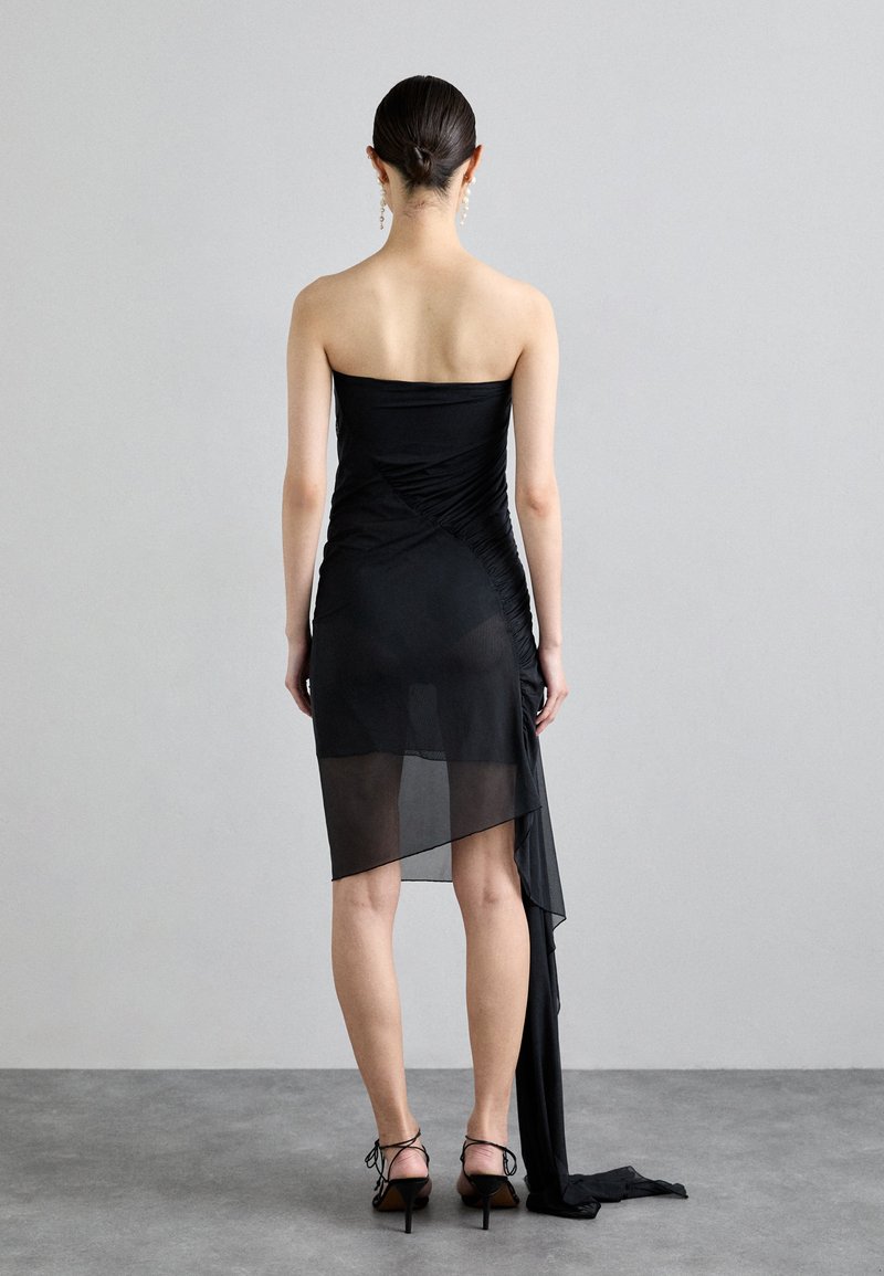 Abito nero senza spalline con texture arricciata, orlo asimmetrico trasparente e drappeggio laterale fluido. Indossato con scarpe con tacco alto nere.
