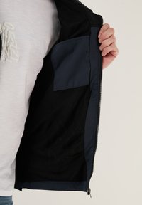 Mornarica zip-up jakna z mrežasto podložjo; notranji žep na levi strani; kontrastne teksture blaga vidne ob odprtju.