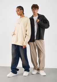 Duas pessoas estão lado a lado. Uma veste um moletom bege e calças de ganga largas azuis; a outra usa uma camisa de couro preta e calças bege.